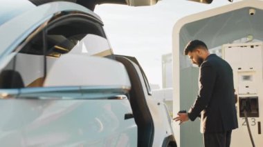 Genç adam elektrikli arabanın yanında dururken gülümsüyor ve kameraya bakıyor. Bir elinde güç kablosu, diğer elinde kahve fincanı tutan mutlu Hintli adam. Ekoloji ve modern yaşam tarzları kavramı
