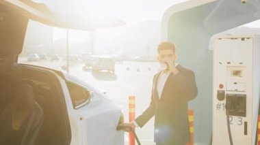 İyi giyimli Arap adam, şehir istasyonunda beyaz elektrikli araba şarj ederken sıcak aromalı kahvenin tadını çıkarıyor. Bilinç ve çevre dostu otomobil kavramı. Çevre taşımacılığı şarj ediliyor