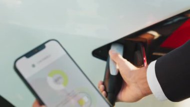 Siyah takım elbiseli bir adam elektrik kablosu takıyor ve mobil ekranı elektrikli araba aküsünün şarj durumuyla gösteriyor. Teknoloji ve yenilik kavramı. Adam arabayı şarj ediyor ve akıllı telefon kullanıyor..