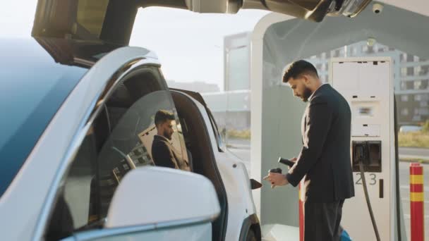 高級電気自動車にプラグインを挿入しながら 片手でコーヒーを保持する正式な衣装でハンサムなアラビア人男性の側面図 現代のエコ輸送の充電プロセス 電気自動車を充電する男 — ストック動画
