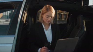 Kafkasyalı iş kadınının modern dizüstü bilgisayarı ve elinde kahve fincanıyla elektrikli arabada oturması. Resmi takım elbiseli çekici bir kadın dışarıda bekliyor.
