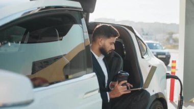 Kamu istasyonunda elektrikli arabayı şarj ederken kablosuz laptop kullanan şık bir girişimci. Uzaklık işleri için modern teknoloji ve yaşam tarzları için ekolojik ulaşım. Elektrikli araba yükleniyor..