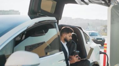 Takım elbiseli, kahve içen ve dizüstü bilgisayarla uğraşan modern elektrikli arabanın pilini şarj eden bir adam. İnsanlar, ulaşım ve teknoloji kavramı. İş adamı arabada otururken.
