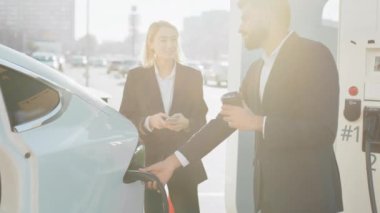 Gülümseyen beyaz kadın ve resmi giyinmiş yakışıklı Hintli adam akıllı telefon kullanıyor ve EV istasyonunda kahve içiyor. İki iş arkadaşı araba şarj ederken iş hakkında konuşuyor.