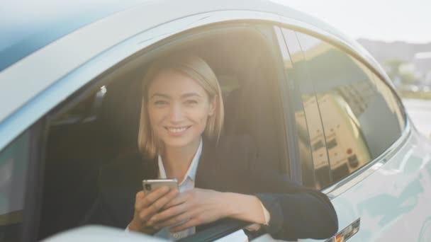 現代的なスマートフォンを手にした魅力的な白人女性が高級車の中に座りながら オートステーションでバッテリーを充電します 出張中にオンラインで働くスタイリッシュなスーツのビジネス女性 — ストック動画