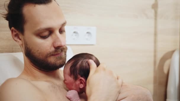 父親は息子と一緒に病院で椅子に座っている 父親の乳房でかわいい新生児の男の子の肖像画を閉じる 妊娠後部の病院で皮膚を剥がす彼の赤ちゃんの世話をする — ストック動画
