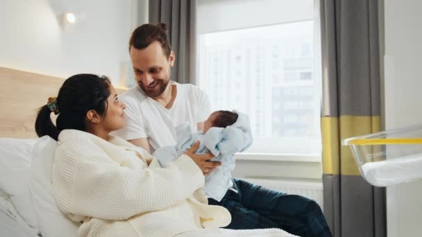 在医院病房里抱着新生儿的快乐父母 笑着和丈夫坐在医院的病床上 抱着婴儿的儿子 爸爸把新生的儿子裹在毯子里送给他的妻子 — 图库视频影像
