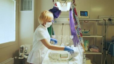 Koruyucu maskeli sarışın kadın doktor yeni doğmuş bebeği kuvözde muayene ediyor. Yenidoğan yoğun bakım ünitesi