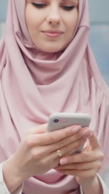 Modern ofis binasının yanında elinde akıllı telefonuyla duran güzel Müslüman kadının alçak açılı görüntüsü. Resmi kıyafet ve tesettürlü genç iş kadını iş e-postalarını kontrol ediyor.