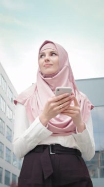 Dikey Ekran: Ofis merkezinde cep telefonu olan Müslüman iş kadını. Modern ofis binasının yanında elinde akıllı telefonuyla duran güzel Müslüman kadının alçak açılı görüntüsü.