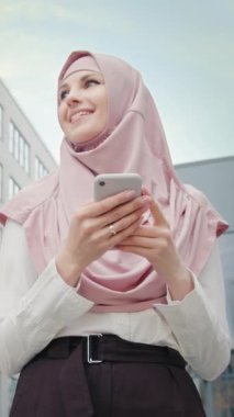 Dikey Ekran: Modern ofis binasının yanında elinde akıllı telefonuyla duran güzel Müslüman kadın. Ofis merkezinde cep telefonu olan Müslüman bir iş kadını. Telefonlu bir kız gülümsüyor..