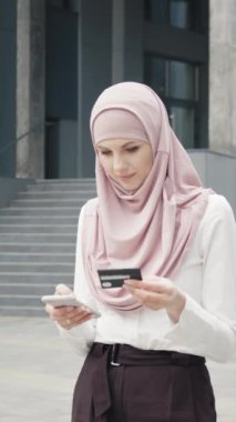 Dikey Ekran: Resmi kıyafetler içindeki güzel Müslüman kadın ve tesettürlü tesettür kredi kartı ve akıllı telefonuyla online alışveriş yapıyor. Genç bayan ofis binasının yanında duruyor ve kolay ödemenin keyfini çıkarıyor.