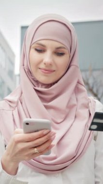 Dikey Ekran: kredi kartı olan Arap kadın. Resmi giyinmiş, tesettürlü güzel Müslüman kadın kredi kartı ve akıllı telefonuyla online alışveriş yapıyor. Ofis binasının yanında duran bayan..