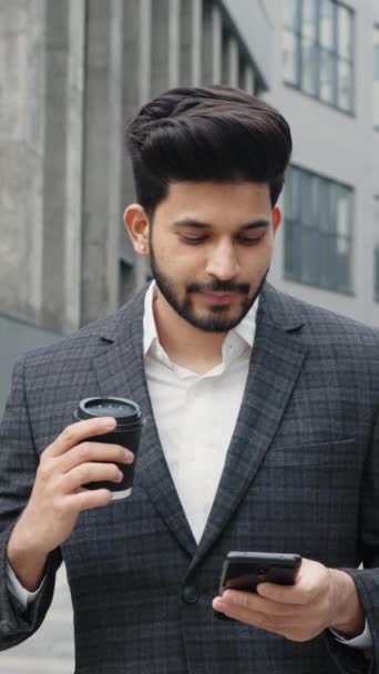 縦スクリーン 現代スマートフォンおよび手のコーヒーのコップが付いているオフィスの建物の近くに立っているビジネスのスーツのアラビア人の男 人々の概念 現代のライフスタイル ブッシー — ストック動画