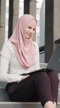 Dikey Ekran: Zarif kıyafetler içindeki çekici Arap kadın portresi ve merdivenlerde dizlerinin üzerinde modern dizüstü bilgisayarla oturan tesettür. Zarif elbiseler ve tesettürlü hoş bir kadın. Müslüman kadın.
