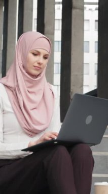 Dikey ekran: şık giyinen ve tesettürlü çekici bir kadın. Uzakta çalışan Müslüman bir kadın. Çekici Arap kadın portresi zarif kıyafetler ve tesettür ile modern merdivenlerde oturan