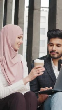 Açık havada dizüstü bilgisayarda çalışan Arap ortaklar. Mutlu Müslüman adam ve kadın açık havada merdivenlerde otururken bir fincan kahveyle kadeh kaldırıyor. İş ortakları.