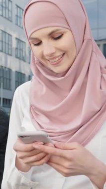 Dikey ekran: tesettürlü hoş bir kadın ahşap bankta oturuyor ve modern cep telefonu tutuyor. Kablosuz dizüstü bilgisayarda çalışan Arap bir adamın bulanık geçmişi. İş kavramı