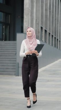Güzel Müslüman kadın elinde akıllı telefonuyla modern ofis binasının yakınına gidiyor. Ofis merkezinin yakınında cep telefonu olan Müslüman bir iş kadını. Kameraya gülümseyen kız.