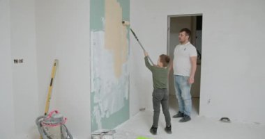 Baba ve oğul bir silindirle bir duvar boyarlar. Apartmanda tamirat yapılıyor. Kendin yap ev onarımı konsepti. Aile zamanı. Slow Motion çekimi. Çocuklar babaya yardım eder, aile onarım projesi