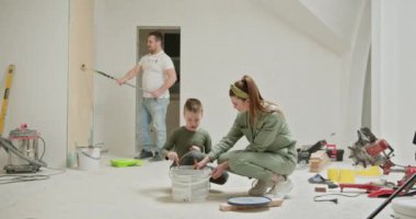 Üç kişilik bir aile, yeni dairelerinin iç duvarlarını boyamaya odaklanan bir ev yenileme projesiyle meşgul. Baba bir silindirle resim yaparken anne ve oğlu yakınlarda boya yapıyor..