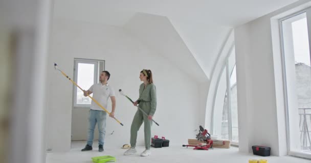 若いカップルは 新しいアパートの壁を描くように家の改良に従事しています 大きな窓付きの広々とした客室は 家族の自己修復の瞬間の背景を作成します — ストック動画