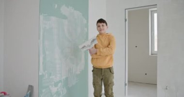 Yeni inşa edilmiş bir dairede, duvarları kısmen boyanmış alçı bir aletle duran genç bir çocuk gururla ayakta duruyor. DIY ev tadilatı ve aile projelerine katkıda bulunmanın sevinci.