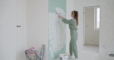 Günlük giysiler içindeki genç bir kadın duvarları beyaza boyayarak yeni dairesini yeniliyor. Özonarım ve ev dekorasyonunun özü özenle boya sürerken, boşluğu yeni bir boyayla değiştiriyor.