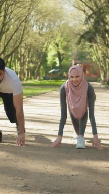 Dikey Ekran: Spor elbiseli genç müslüman çift yeşil yaz parkında koşmak için başlangıç pozisyonunda. Erkek ve kadın tesettürde sabah sporuna hazırlanıyor.