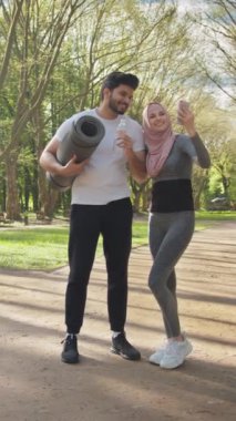 Dikey Ekran: Spor elbiseli genç müslüman çift açık hava aktivitesinden sonra selfie çekmek için modern akıllı telefon kullanıyor. Elinde su ve yoga minderi tutan bir adam, elinde cep telefonu tutan bir kadın..