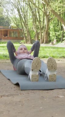 Dikey ekran: tesettürlü ve spor elbiseli hoş bir kadın yoga minderinde oturuyor ve vücudunu esnetiyor. Genç Müslüman kadın eğitimi sabah vakti açık havada. Müslüman kadın yoga minderinde eğitim görüyor.