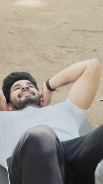 Dikey Ekran: Kas Hindu adam ayaklarını tutan tesettürlü kadının yardımıyla karın kaslarını yoga minderinde çalıştırıyor. Aktif genç çift temiz havada birlikte antrenman yapıyor. Hintli adam karın kası çalıştırıyor.