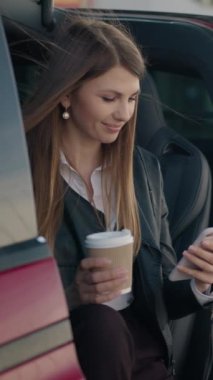Dikey Ekran: Portre Çekici kadın içeride oturuyor modern akıllı telefon ve elinde bir fincan kahve ile elektrikli araba şarj ediyor. Genç modern kız telefondan kameraya bakar ve gülümser.