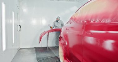 Koruyucu kıyafet giyen bir araba boyacısı servis odasındaki bir araba tamponuna kırmızı boya sürüyor. Otomotiv boyama ve kaporta tamiri devam ediyor. Araba üretim fabrikası