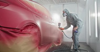 Koruyucu takım elbiseli ve maskeli bir araba boyacısı araba tamirhanesinde sprey boyayla araba boyuyor. Otomatik boyacı sprey boya kabinine boya sürüyor. Otomotiv boyama hizmetleri kavramı
