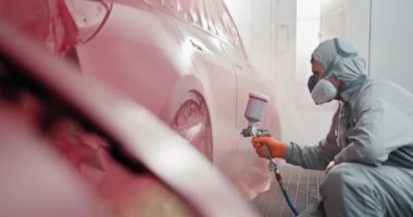 Koruyucu giysili bir araba boyacısı araba bakım odasındaki bir araca araba tamiratı için kırmızı boya uyguluyor. Araba boyacısı oto tamirhanesinde boya uyguluyor. Otomotiv Boyama Servisi