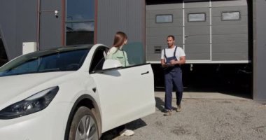 Zarif bayan müşteri, otomobil tamir atölyesinin önünde erkek bir otomotiv teknisyeniyle araba sorunları hakkında konuşuyor. Kadın beyaz elektrikli arabasının yanında duruyor. Kadın ve Tamirci