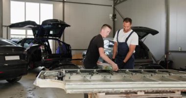 Modern bir oto tamir atölyesinde elektrikli araba akü modülünü tamir eden ve sorun çıkaran iki erkek teknisyen. Atölye Ayarları Çoklu Parçalanmış Araçları içerir. Otomatik Servis
