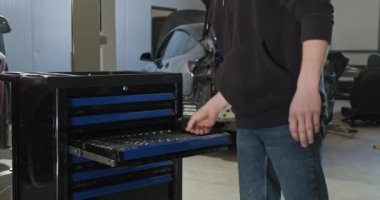 Tamirci bir otomotiv tamirhanesinden alet dolabına giriyor. Arka planda kısmen parçalanmış elektrikli araba, EV bakım ve onarımı vurguluyor. EV onarım konsepti. Otomatik Servis.