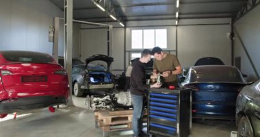 EV onarım konsepti. Otomobil servisi. İki erkek tamirci modern bir otomobil atölyesinde elektrikli araba motoru tamirinde birlikte çalışıyorlar. Arka planda görülen özelleştirilmiş araçlar ve elektrikli araçlar