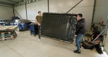 Modern bir atölyede elektrikli araba aküsü modülünü kaldıran ve tamir eden iki teknisyen. Otomotiv endüstrisi, elektrikli araçlar ve pil teknolojisi kavramları. Lityum iyon Bataryası