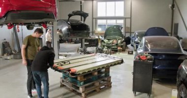 İki teknisyen meşgul bir oto tamir atölyesinde elektrikli araba aküsü modülü üzerinde çalışıyor. Çevredeki araçlar ve ekipmanlar elektrikli araç bakımına odaklandığını gösteriyor. Pil hücre modülü paketi.