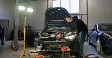 İyi aydınlatılmış bir garajda arabanın elektrik sistemini tamir eden yetenekli bir tamirci. Tamirci araç üzerinde çalışırken çeşitli araç ve gereçler kullanır. Otomatik servis ve bakım kavramı.