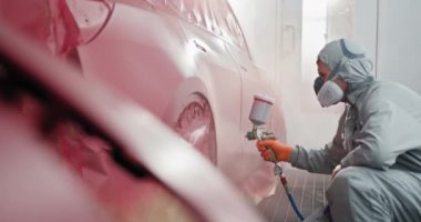 Koruyucu giysili bir araba boyacısı araba bakım odasındaki bir araca araba tamiratı için kırmızı boya uyguluyor. Araba boyacısı oto tamirhanesinde boya uyguluyor. Otomotiv Boyama Servisi