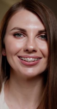 Dikey video. Ağız temizliği ve sağlık hizmetleri. Diş teli takan kadın evde diş fırçası tutarken gülümsüyor. Diş tellerini düzgün bir fırçalama tekniğiyle koruyan bir kadın. Parantezler.