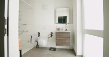 Tekerlekli sandalyeyle yıkanabilen banyo ve modern gösteriş. Beyaz fayanslar ve doğal ışık, temiz ve parlak bir alan yaratır. Tasarım kullanım kolaylığı için erişilebilir özellikleri içerir.