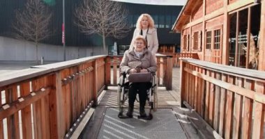 Güneşli bir günde arka planda binalar olan tahta rampada tekerlekli sandalyeli yaşlı bir kadını iten bir kadın. Tekerlekli sandalyedeki yaşlı kadın ve bakıcı şehir meydanını işaret ediyor.