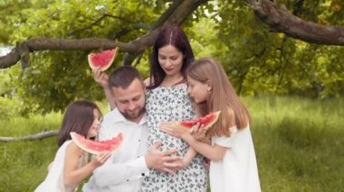Yeşil bahçede birlikte poz verip tatlı karpuz yiyen yaz kıyafetleri içinde beyaz bir aile. Yakışıklı bir adam ve iki tatlı çocuk hamile kadını kucaklıyor. İki çocuklu hamile bir kadın..