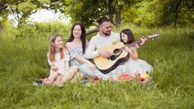 Handsome man playing guitar for his charming wife and two cute daughters. Happy caucasian family sitting on soft blanket at garden and singing songs. Parents with two kids playing guitar on fresh air.