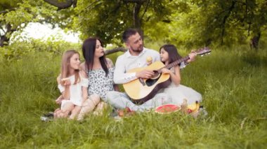 Happy parents with two child sitting on checkered blanket and singing songs during picnic time. Father playing guitar, mother hugging cute daughters. Man playing on guitar for his wife and daughters.